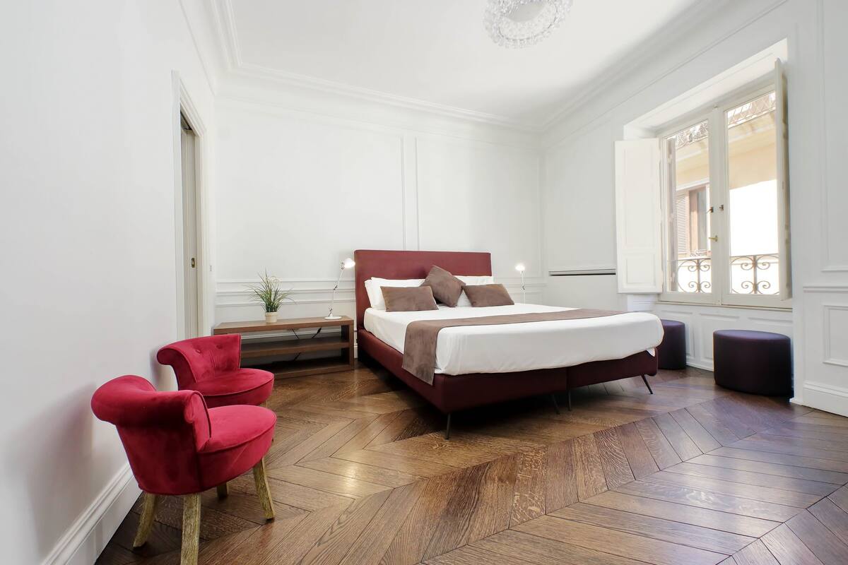 A bedroom with a wooden herringbone floor, a red upholstered chair, a bed with a white and brown headboard, and a window with decorative iron railings.