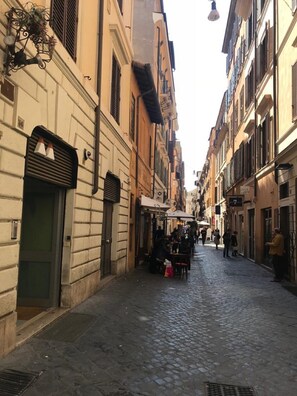 Une ruelle étroite pavée, bordée de bâtiments historiques, dont certains sont dotés de fenêtres à volets et d’espaces aménagés pour s’asseoir à l’extérieur.
