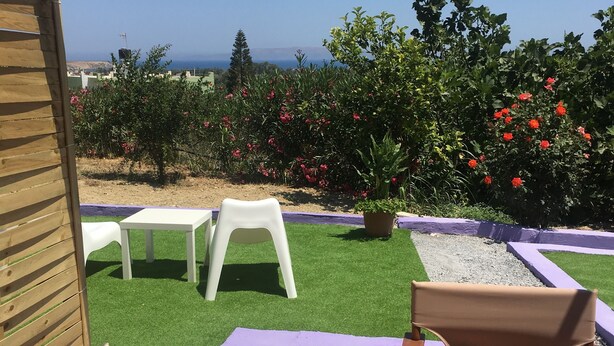 Un área de estar al aire libre con una mesa y sillas de madera, una silla blanca y un mantel morado. Hay plantas en macetas y se ve el mar al fondo.