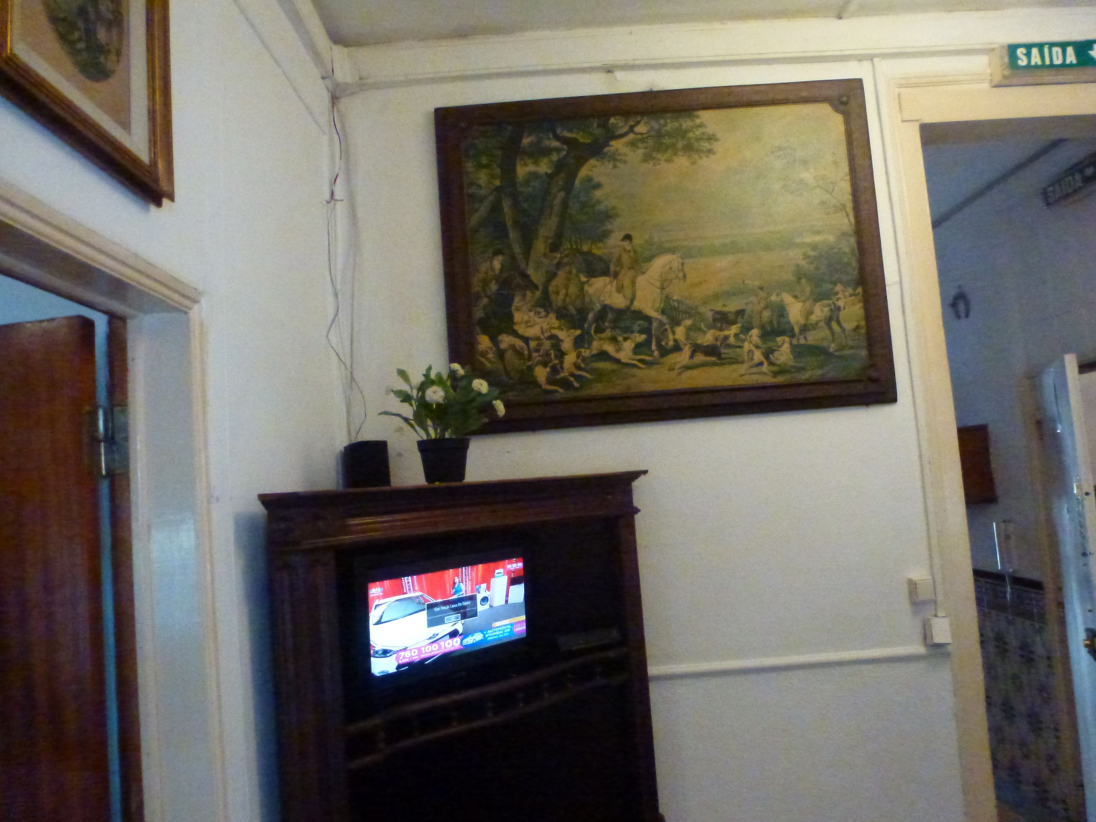 A room with a television on a wooden stand, a framed painting above it, and a door labeled "SAÍDA".