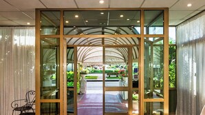 A hotel entrance with a glass door, a tiled floor, and a view of the garden outside. - Hotel Ambasciatori (Pietrasanta)