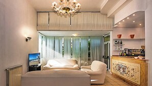 A modern hotel room with a large bed, a sofa, a television, a kitchen area, and a view of the outdoors. - Hotel Ambasciatori (Pietrasanta)
