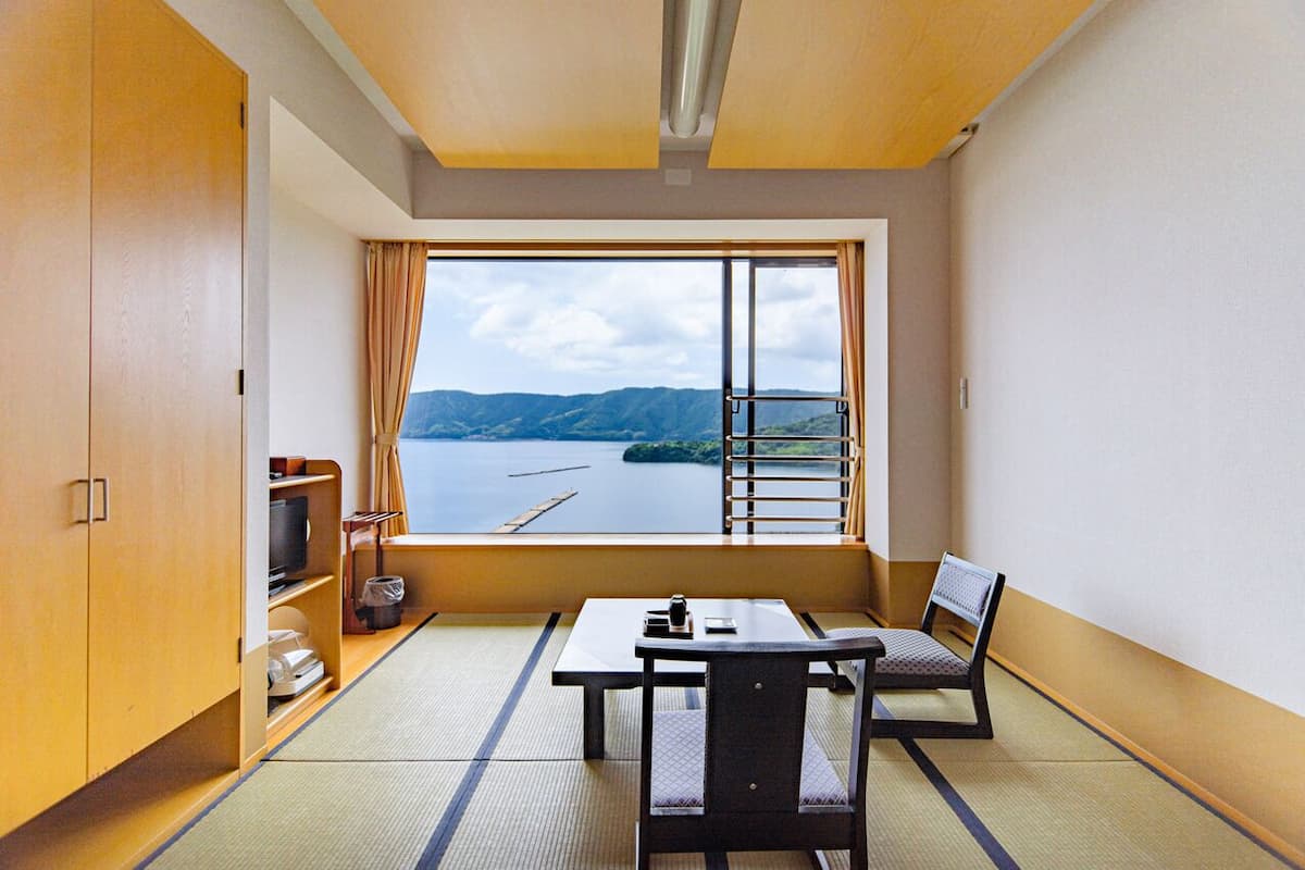 A room with tatami flooring, a table, chairs, a TV, and a window with a view of a lake and mountains.