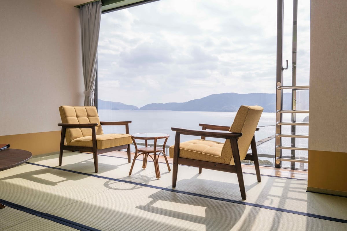 A room with tatami flooring, two wooden chairs, a small round table, and a view of a lake through a window.
