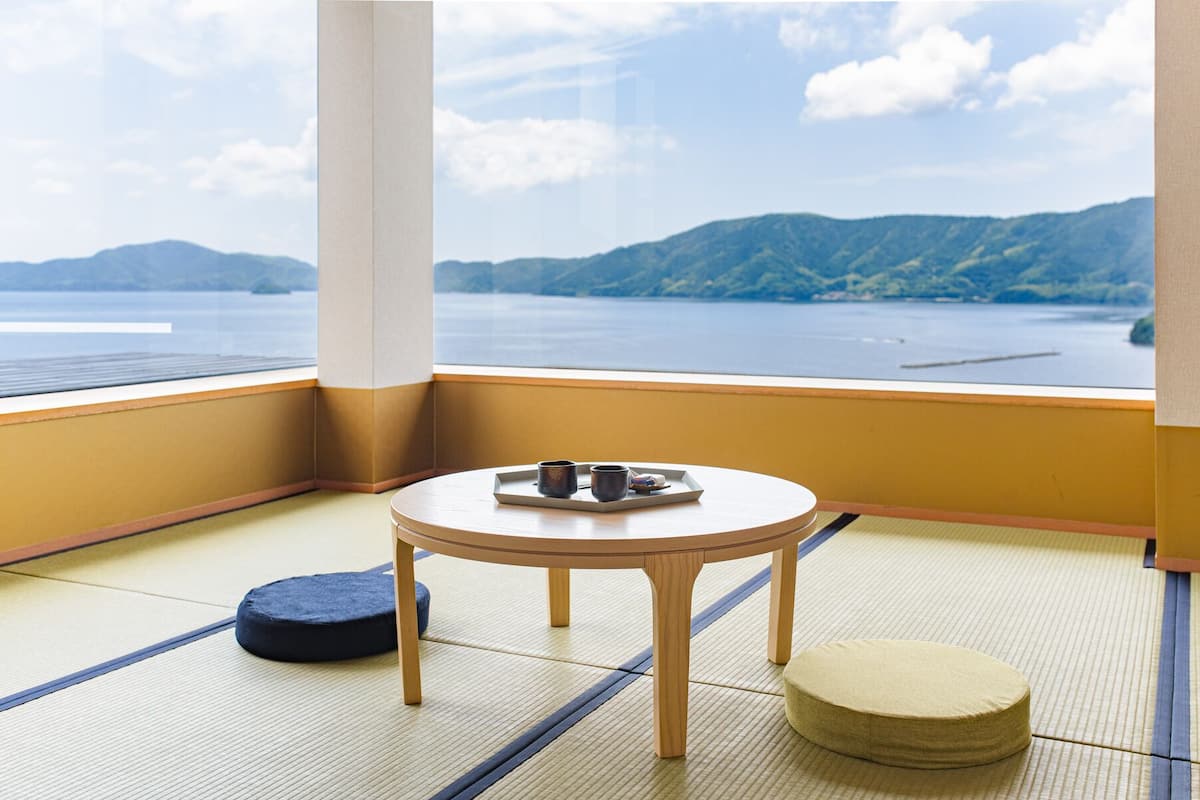 A room with tatami flooring, a round table, and a view of a lake and mountains.