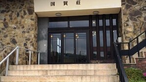 A building entrance with a stone facade, a set of stairs leading up to a dark-colored door, and a sign above the door. - Kunigaso (Nishinoshima)