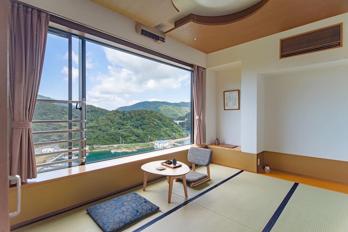 A room with a large window, tatami mat flooring, a small table, a chair, and a view of a river and mountains.