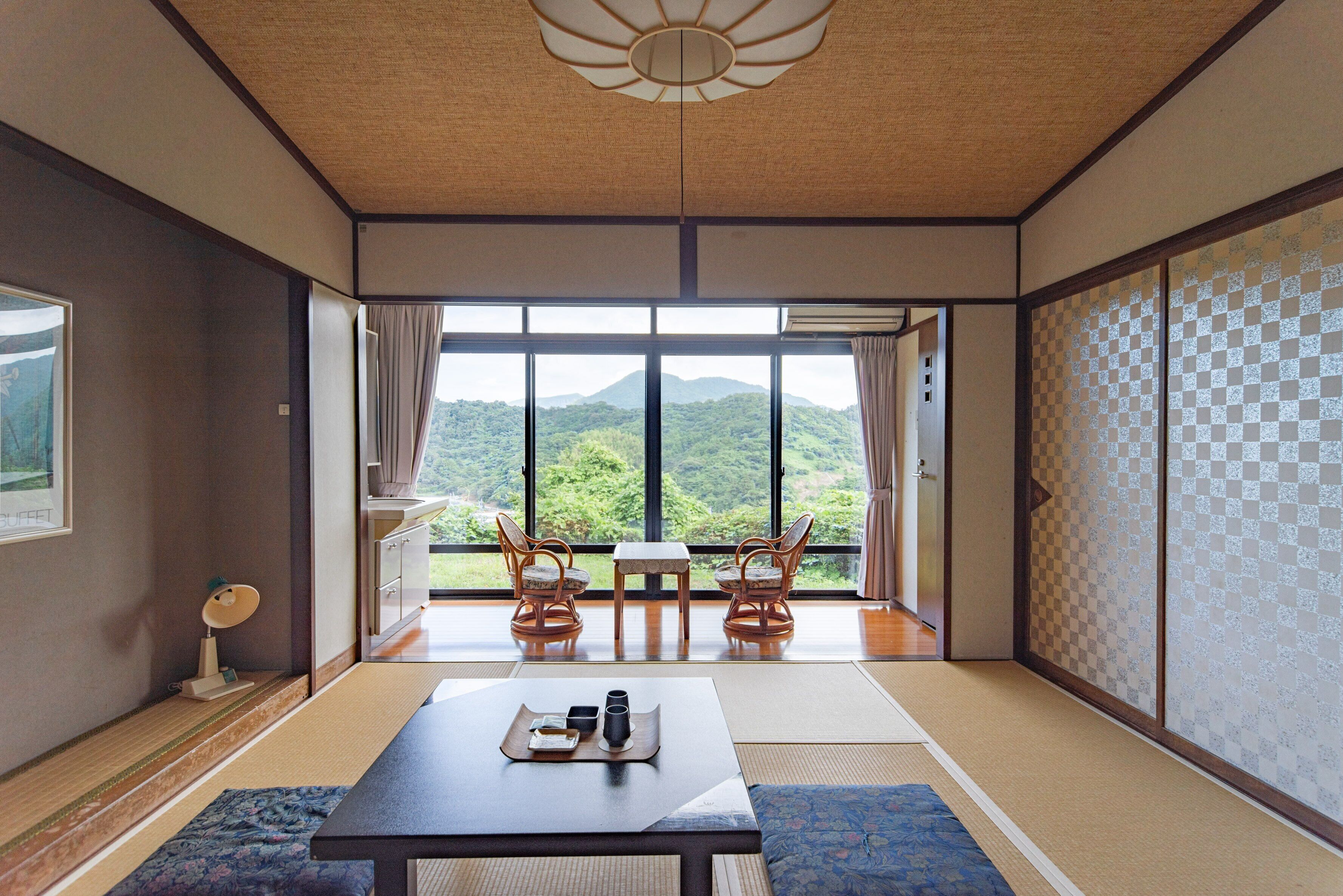 Una habitación japonesa tradicional con suelo de tatami, una mesa baja con bandeja, dos sillas y vista a una montaña a través de amplios ventanales.