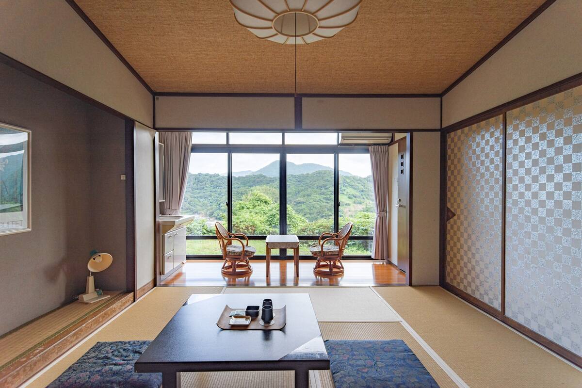 A traditional Japanese room with tatami flooring, a low table with a tray, two chairs, and a view of a mountain through large windows.