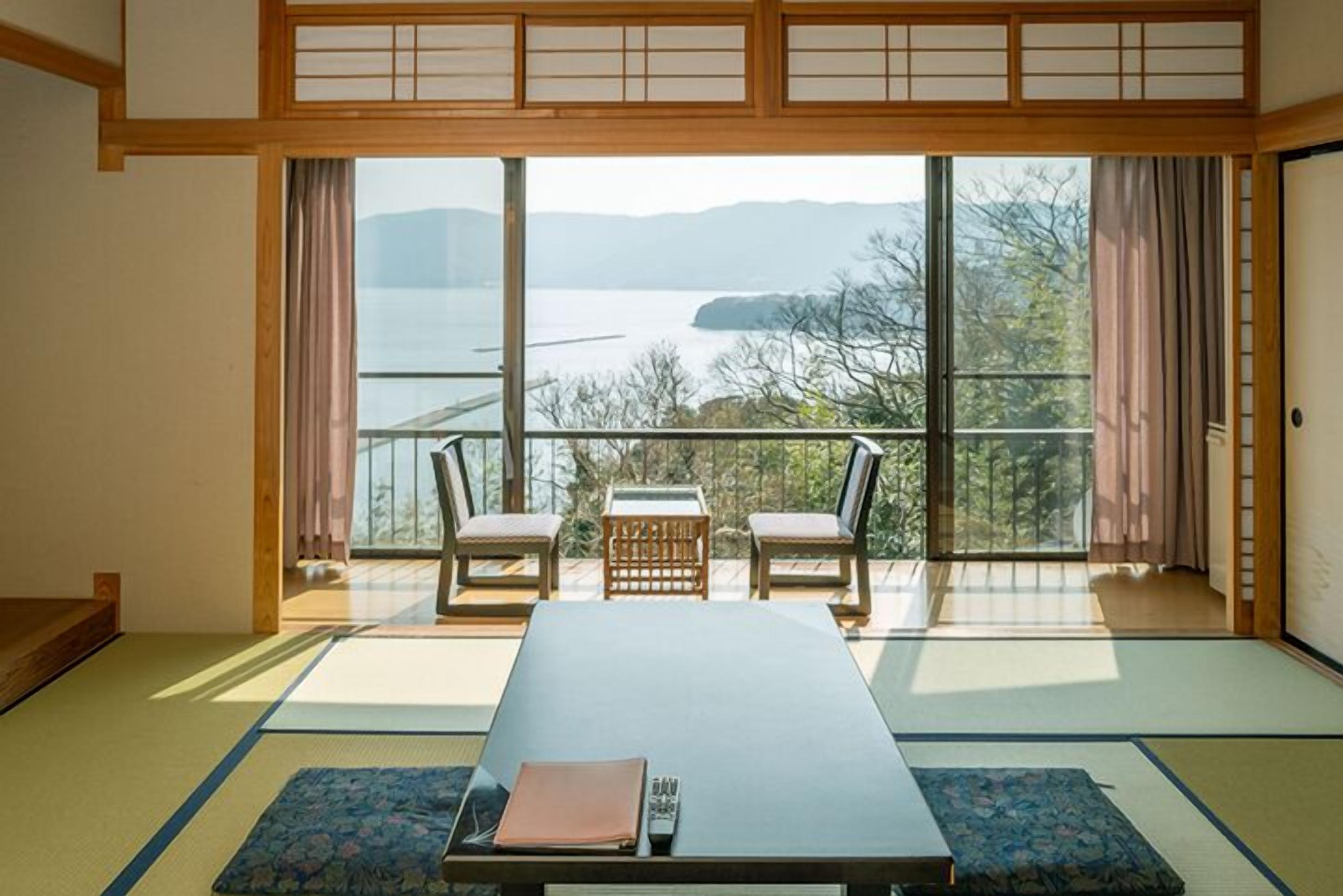 Una habitación japonesa tradicional con suelo de tatami, una mesa baja, dos sillas y vistas a un lago a través de amplias ventanas.