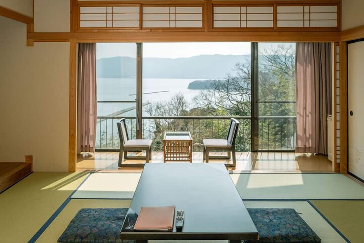 A traditional Japanese room with tatami flooring, a low table, two chairs, and a view of a lake through large windows.