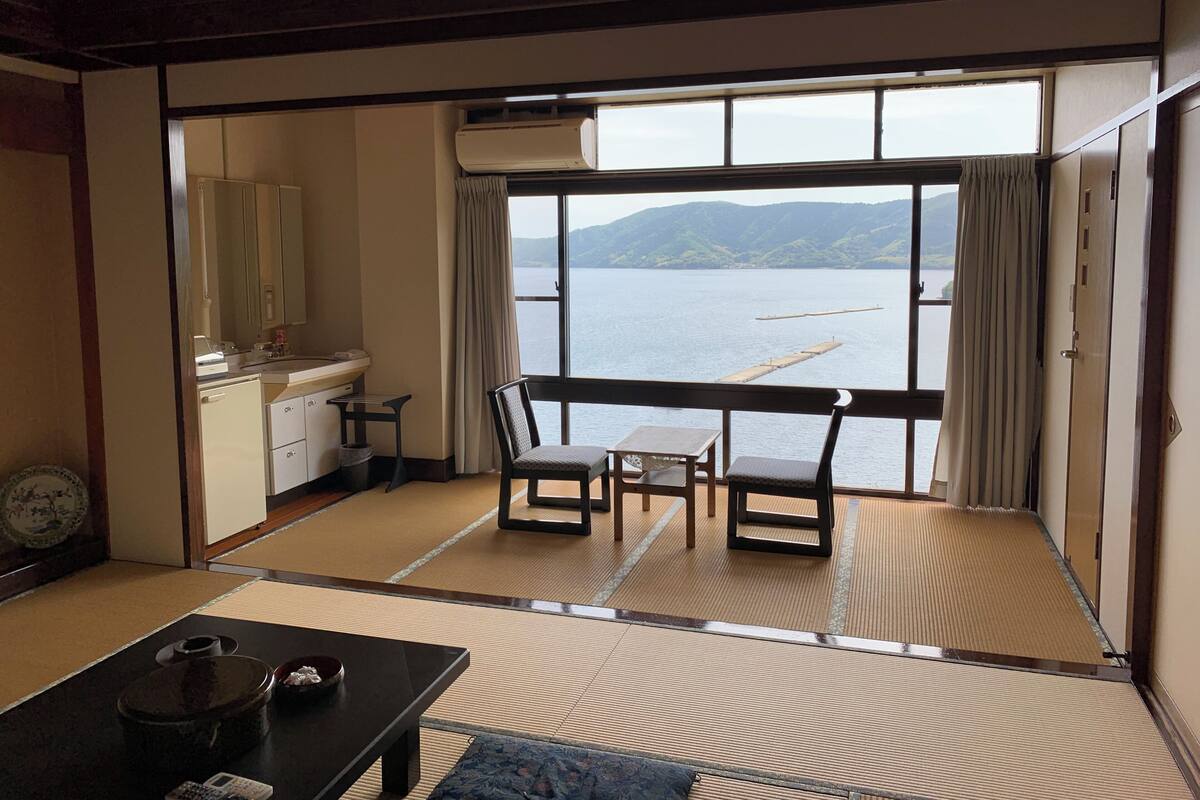 A room with tatami flooring, a desk, a chair, and a view of a lake with mountains in the distance.