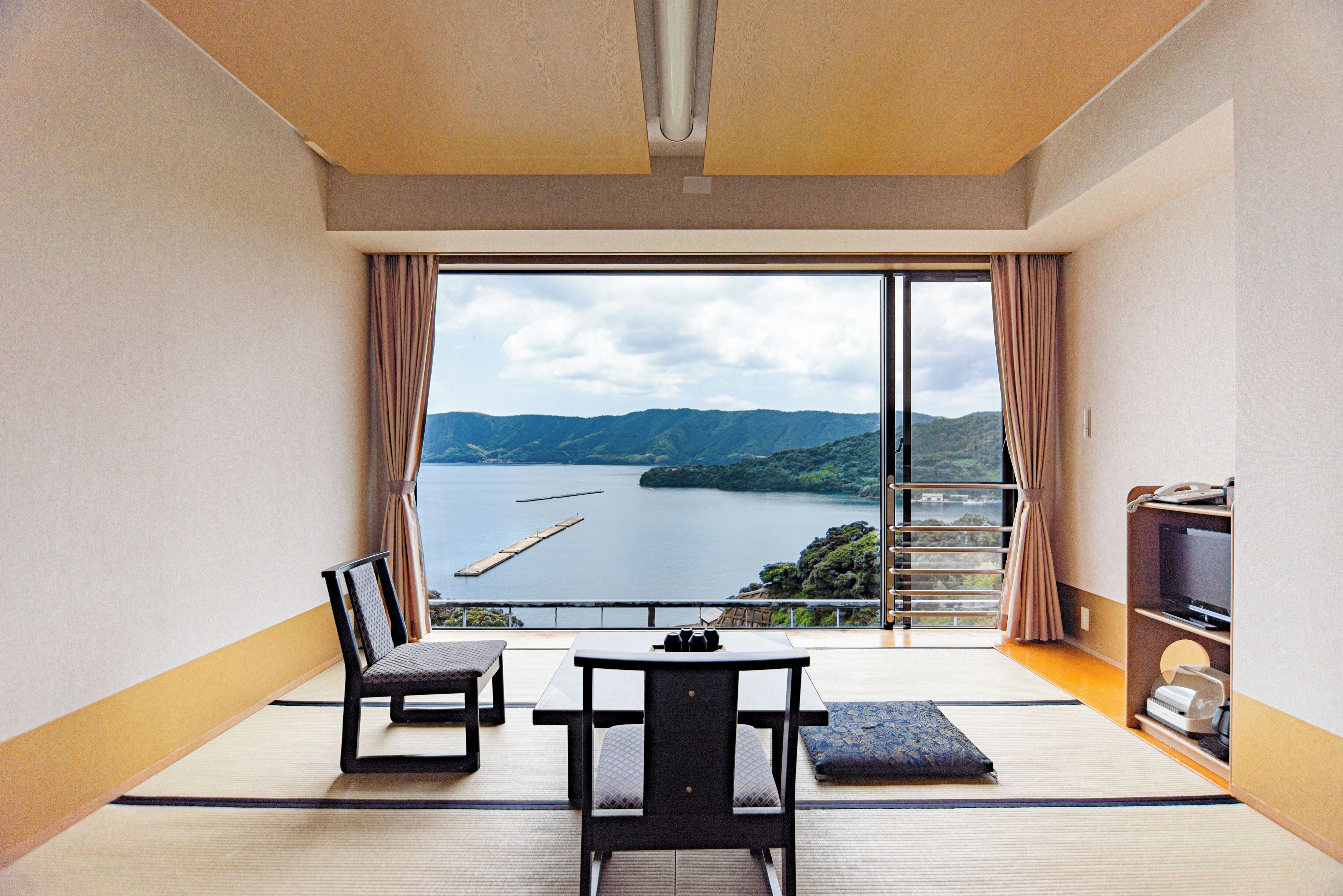 Una habitación con suelo de tatami, una mesita baja, dos sillas, un televisor y un ventanal con vistas a un lago y montañas.
