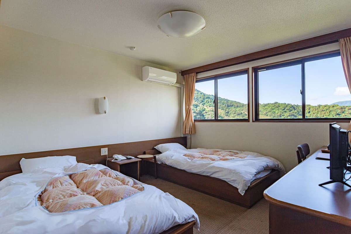 A hotel room with two beds, a desk, a computer, and a window with a view of green hills.