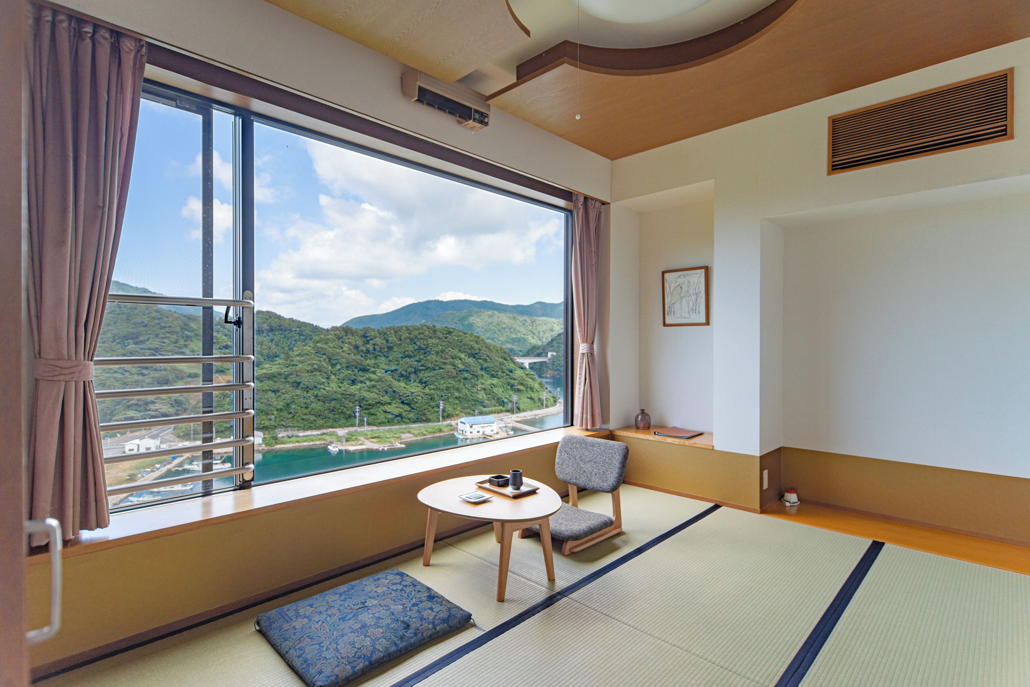 Una habitación con suelo de tatami, vista a un río y montañas, una mesita con una taza, y una silla.