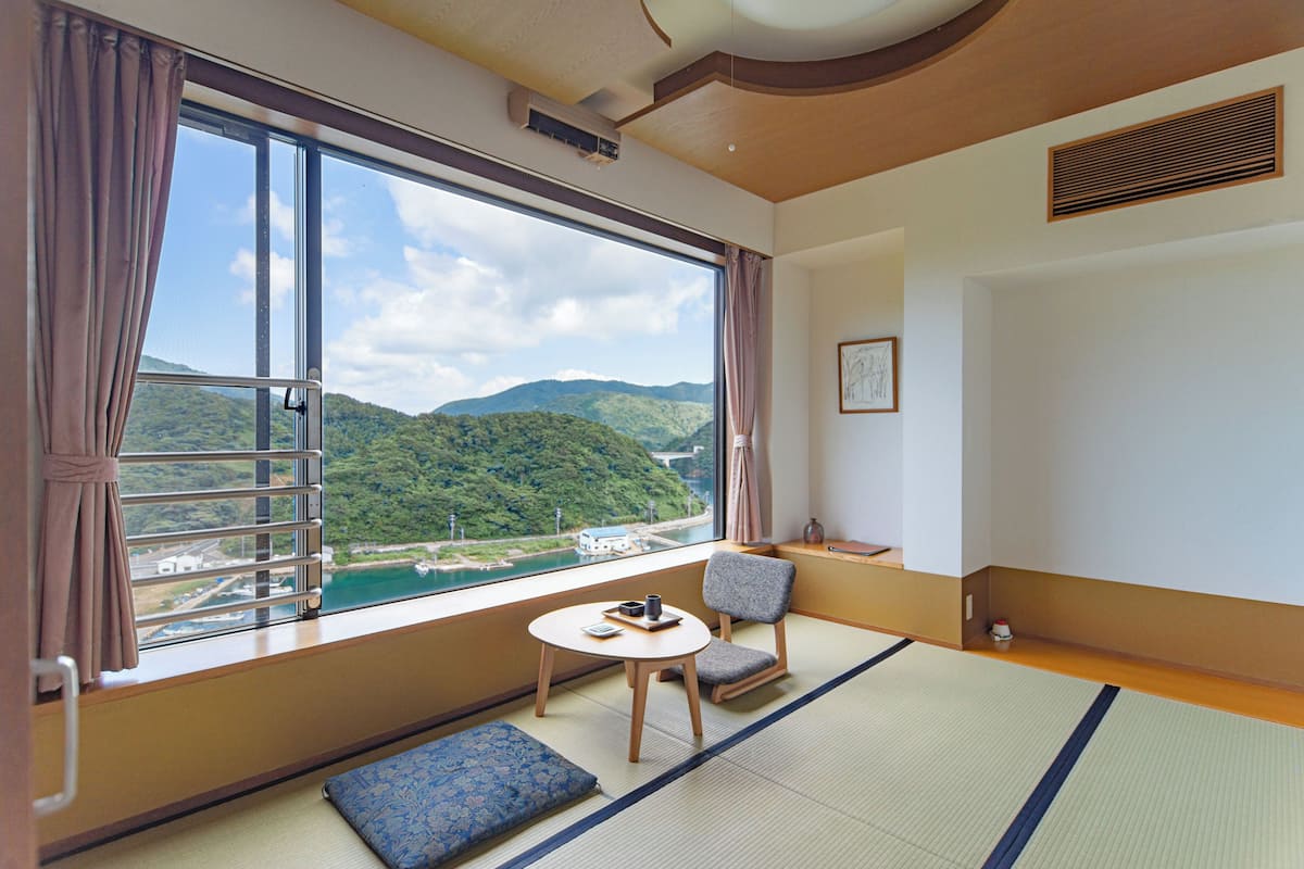 A room with tatami flooring, a view of a river and mountains, a small table with a cup, and a chair.