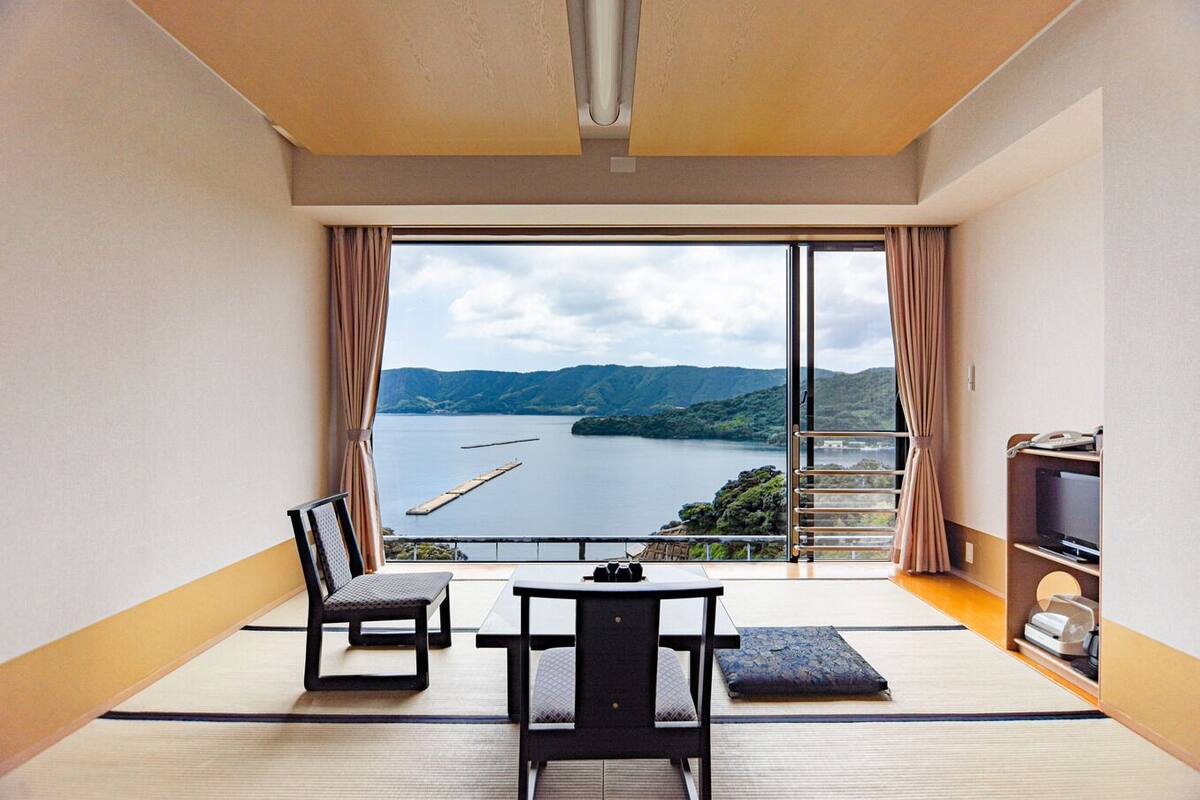A room with a large window, a table, two chairs, a TV, and a small shelf with items.