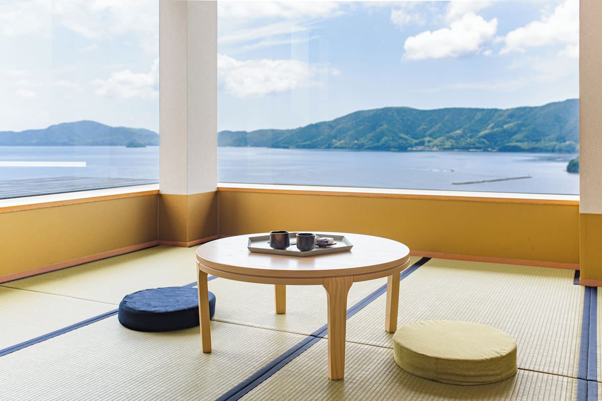 A tatami mat room with a view of a lake and mountains, a small round table with a tray, and two cushions.