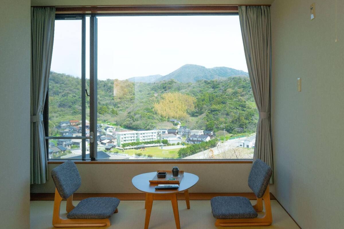 A room with a large window, two chairs, a small table, and a view of a residential area with houses and mountains.