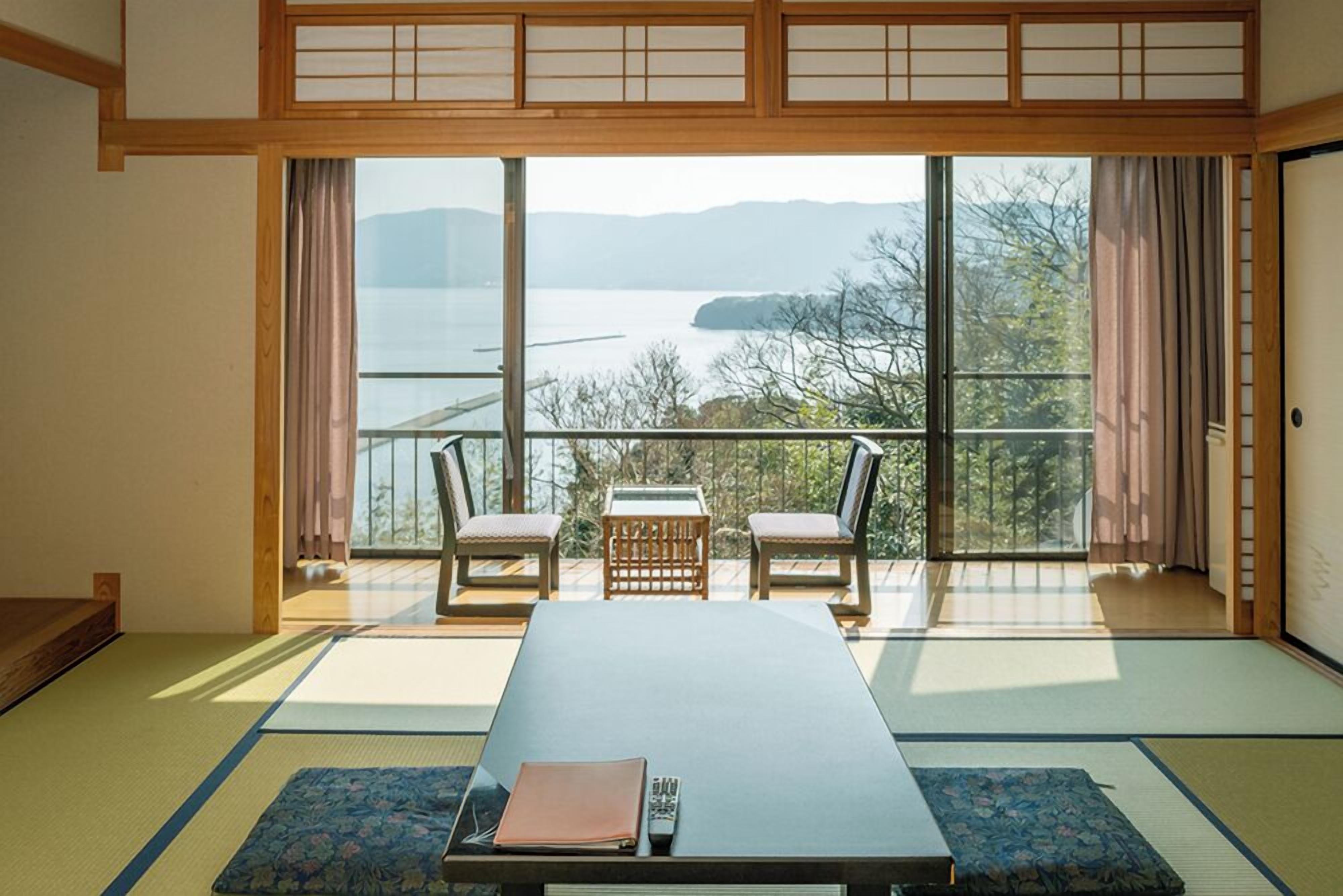 Una habitación japonesa tradicional con suelo de tatami, una mesa baja, dos sillas y vistas a un lago a través de amplias ventanas.