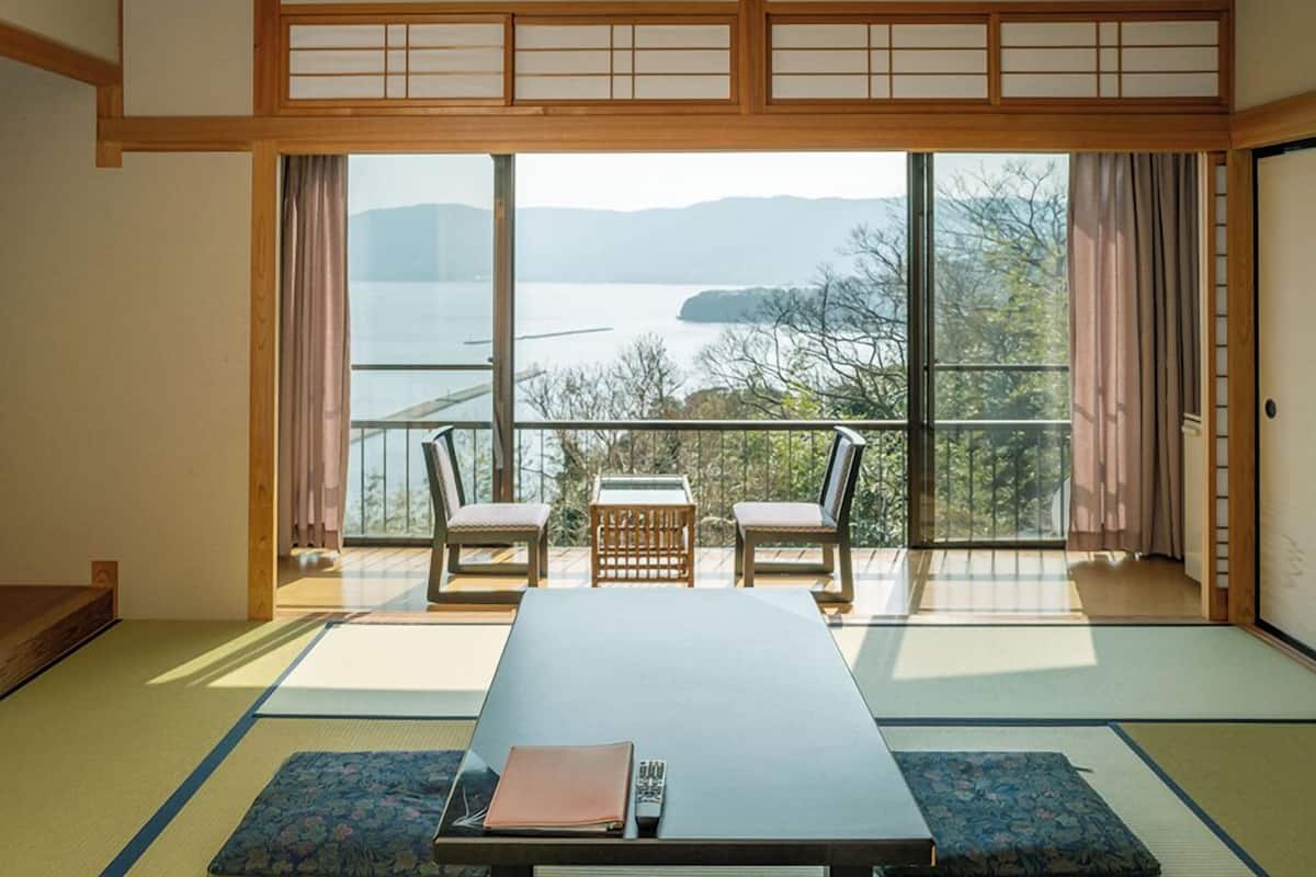 A traditional Japanese room with tatami flooring, a low table, two chairs, and a view of a lake through large windows.