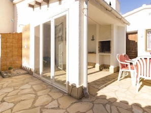 Un espace extérieur aménagé en patio, avec un sol en pierre, une porte vitrée donnant sur l’intérieur et une table entourée de chaises.