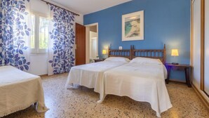 A room with two beds, a large window with blue curtains, a painting on the wall, and a sun-shaped ceiling light.