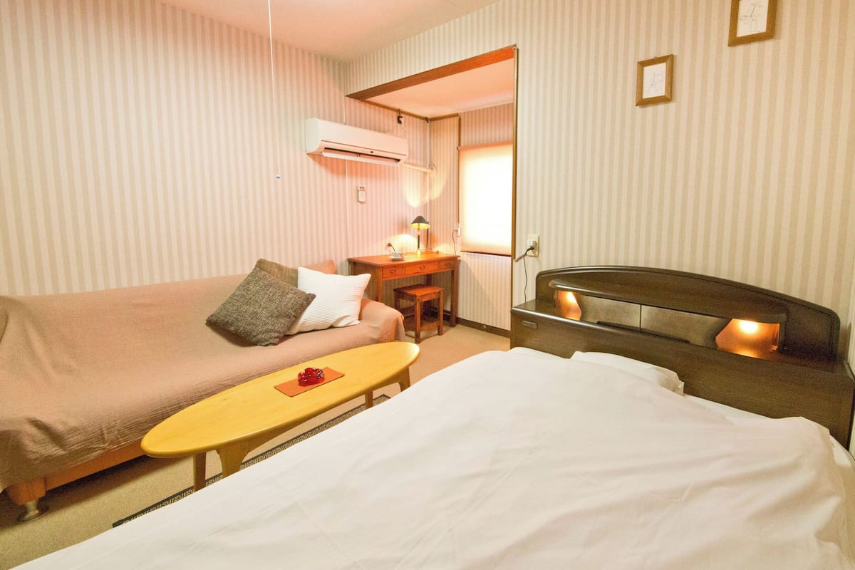 A hotel room with a bed, a sofa, a desk, and an air conditioning unit.