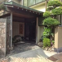 Una entrada tradicional japonesa con muro de piedra, galería techada y un pequeño jardín con un bonsái.