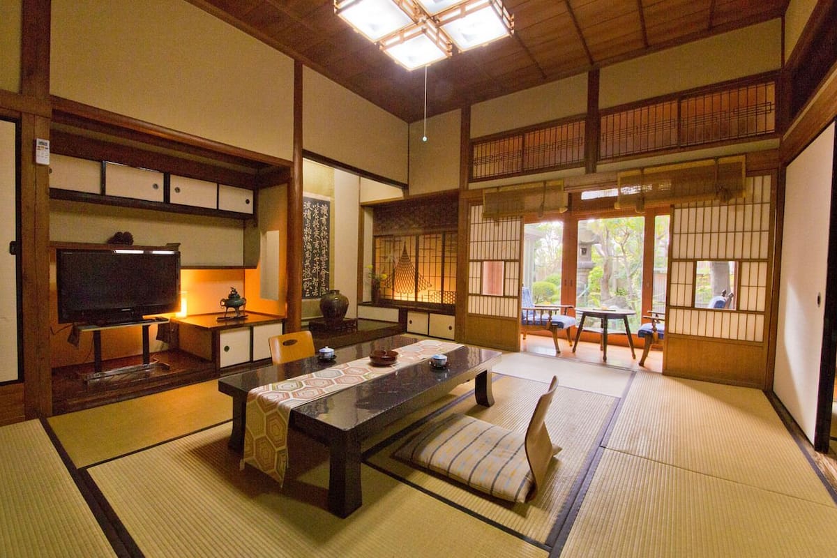 A traditional Japanese room with tatami flooring, a low table with cushions, a television, and sliding doors leading to a balcony with a view of greenery.