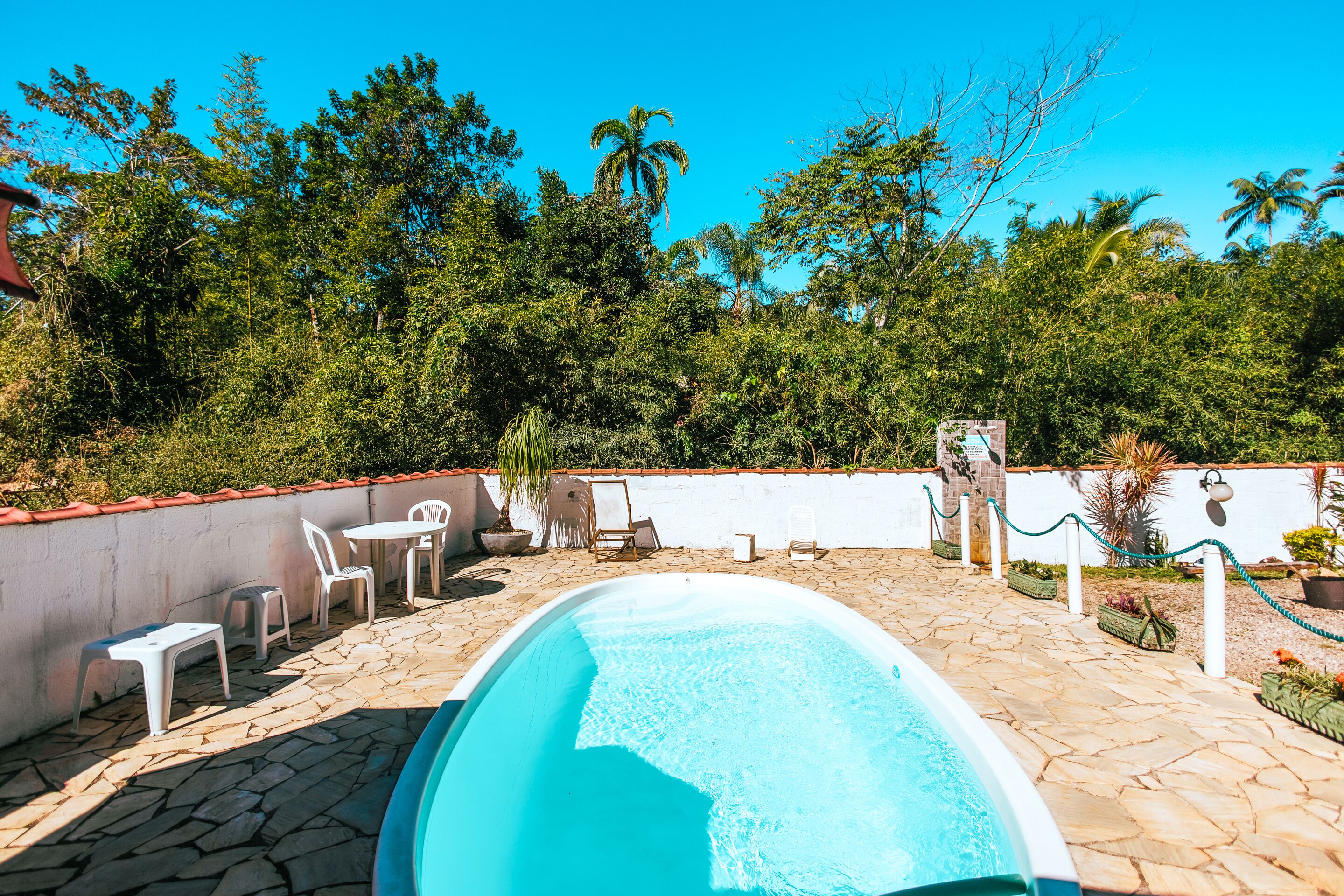 Une piscine avec une terrasse en pierre, entourée de chaises et de tables blanches, et un mur blanc avec un palmier en arrière-plan.