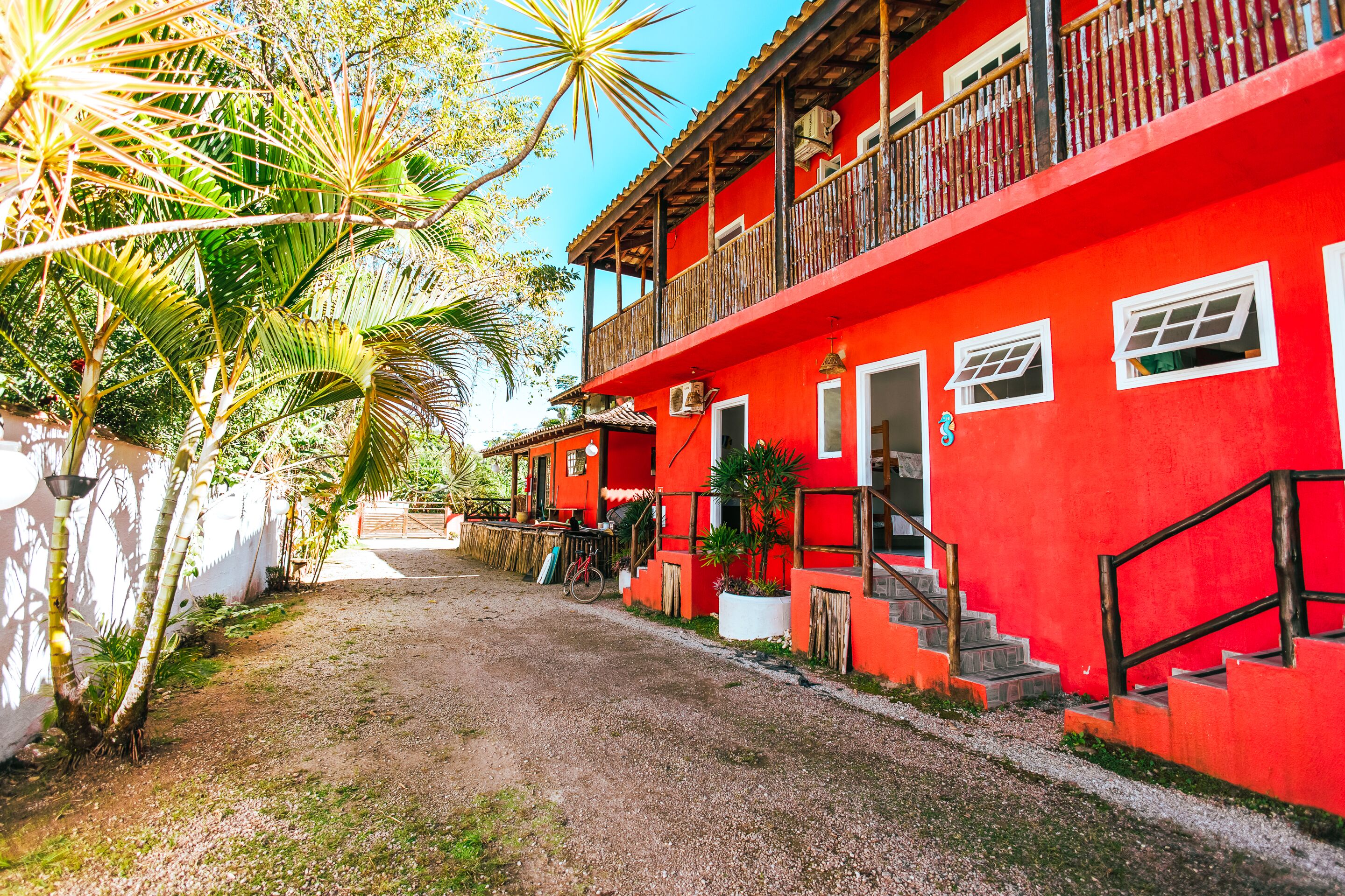 Um edifício vermelho com varanda, escadaria e um caminho de terra.