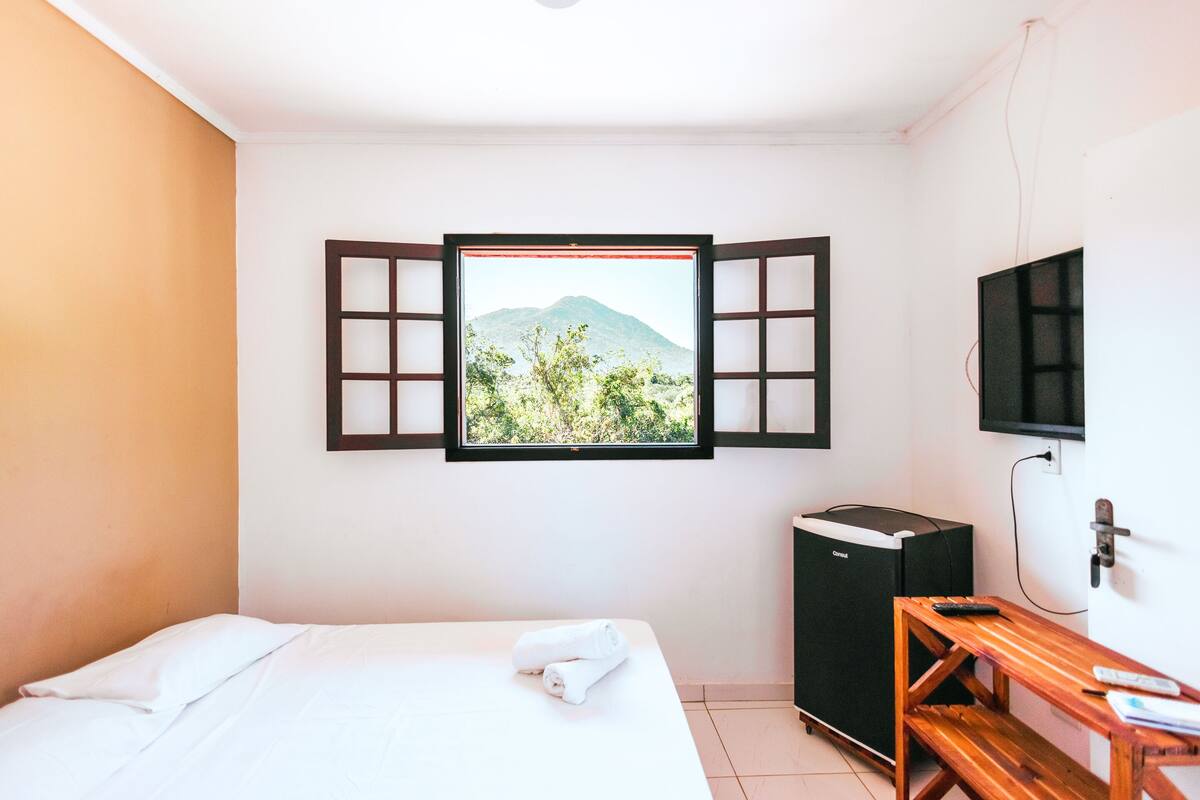 A hotel room with a bed, a wooden bench, a TV, and a window with a view of a mountain and greenery.