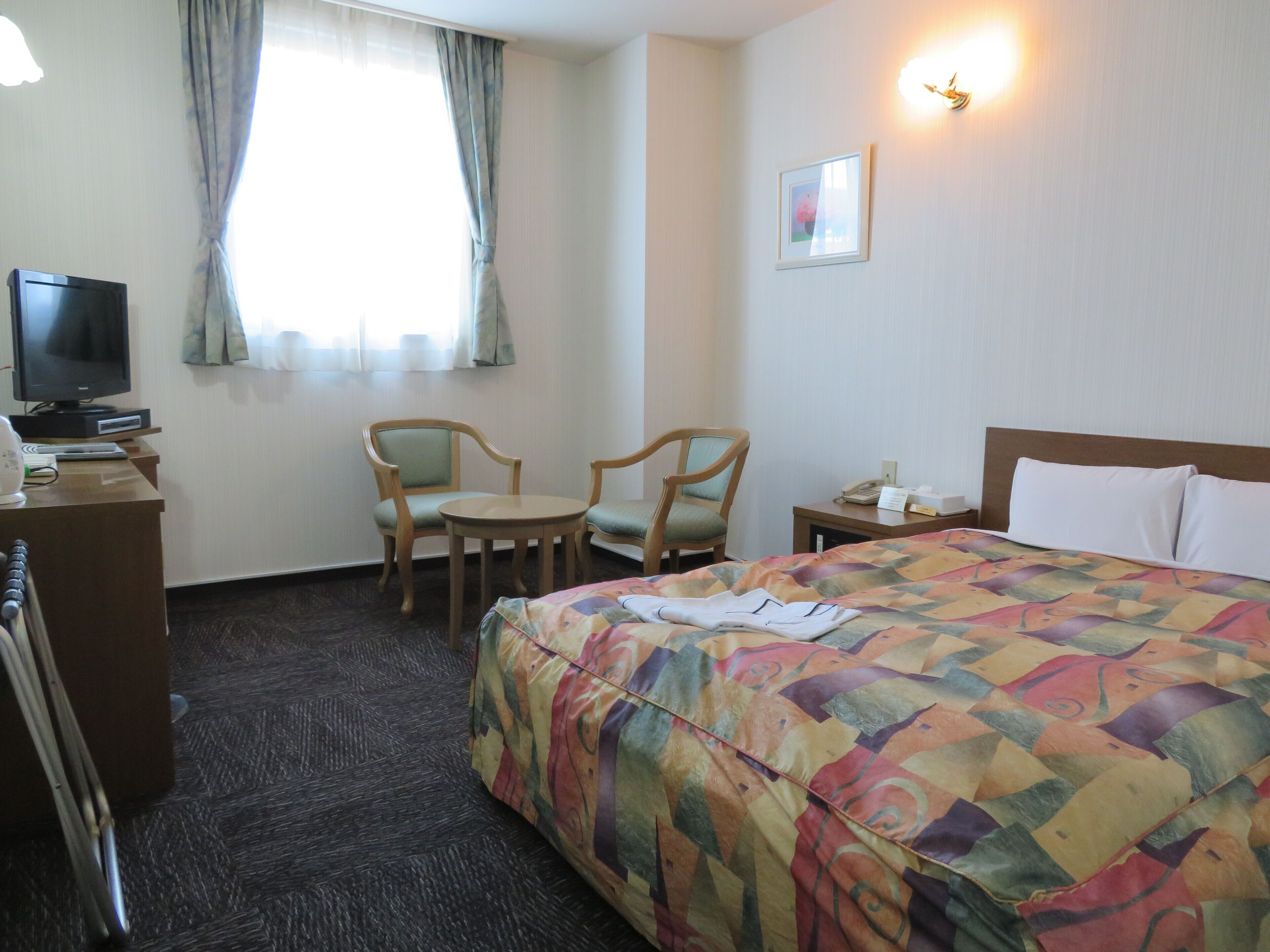 A hotel room with a bed, two chairs, a desk with a television, and a bedside table with a lamp.