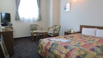 A hotel room with a bed, two chairs, a desk with a television, and a bedside table with a lamp.