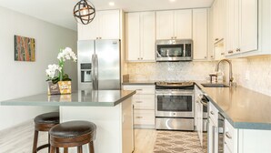 A modern kitchen with stainless steel appliances, a herringbone tile backsplash, and a central island with bar stools. - Kihei Wailea Hale across from World Famous Keawakapu Beach! (Kihei)