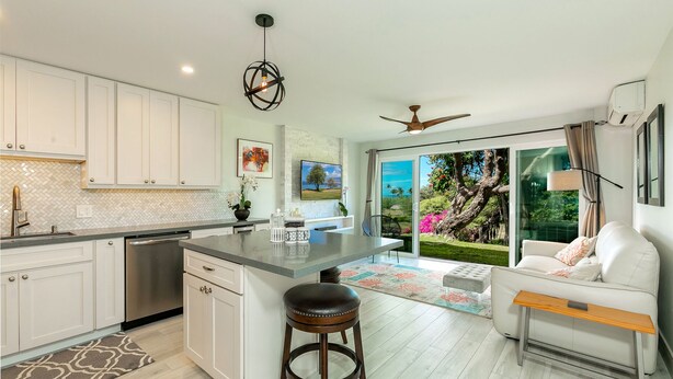 A modern kitchen with white cabinets, a central island, and a dining area with a white sofa and wooden side table. - Kihei Wailea Hale across from World Famous Keawakapu Beach! (Kihei)