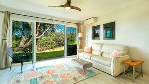 A modern living room with a sliding glass door leading to a garden, a white sofa, a small table, and a ceiling fan. - Kihei Wailea Hale across from World Famous Keawakapu Beach! (Kihei)