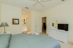 A bedroom with a bed, a dresser, a television, a ceiling fan, and a bathroom visible through an open door. - 14543 Abaco Lakes Dr #104 (Cape Coral)
