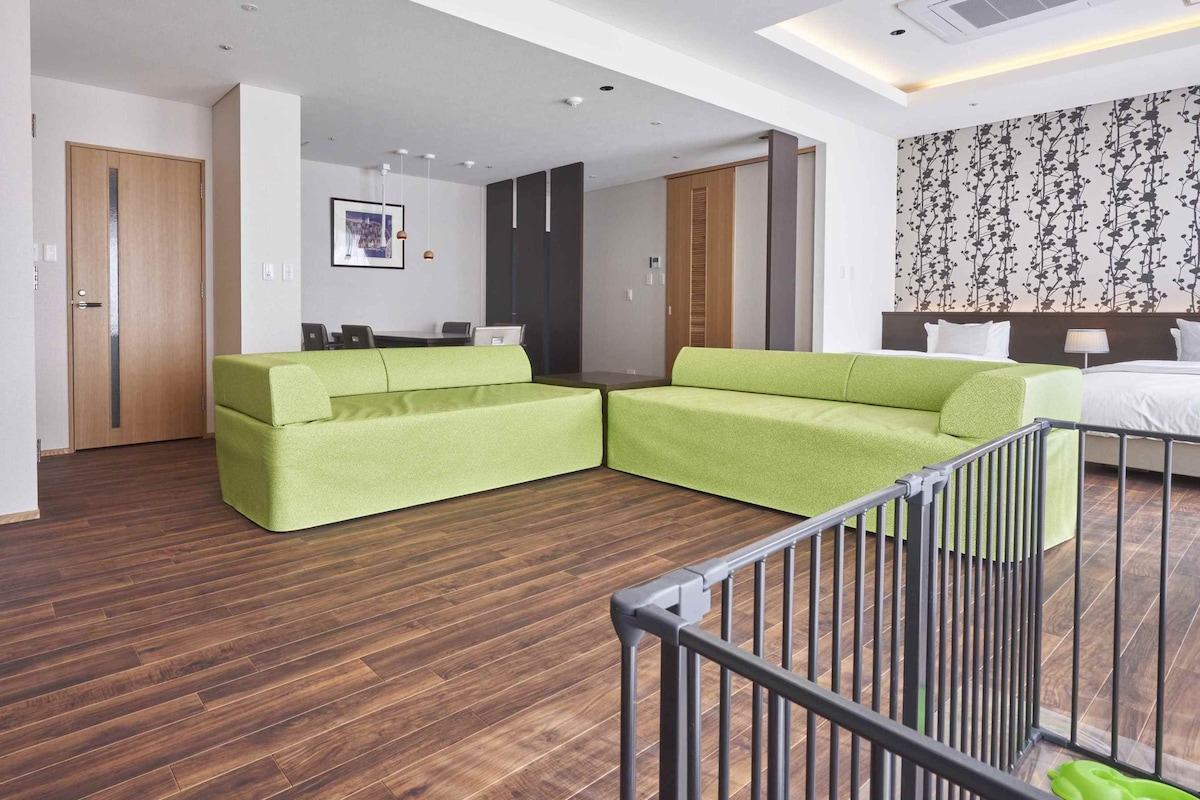 Una habitación de hotel moderna con piso de madera, un sofá verde, una cama y un escritorio con una silla.