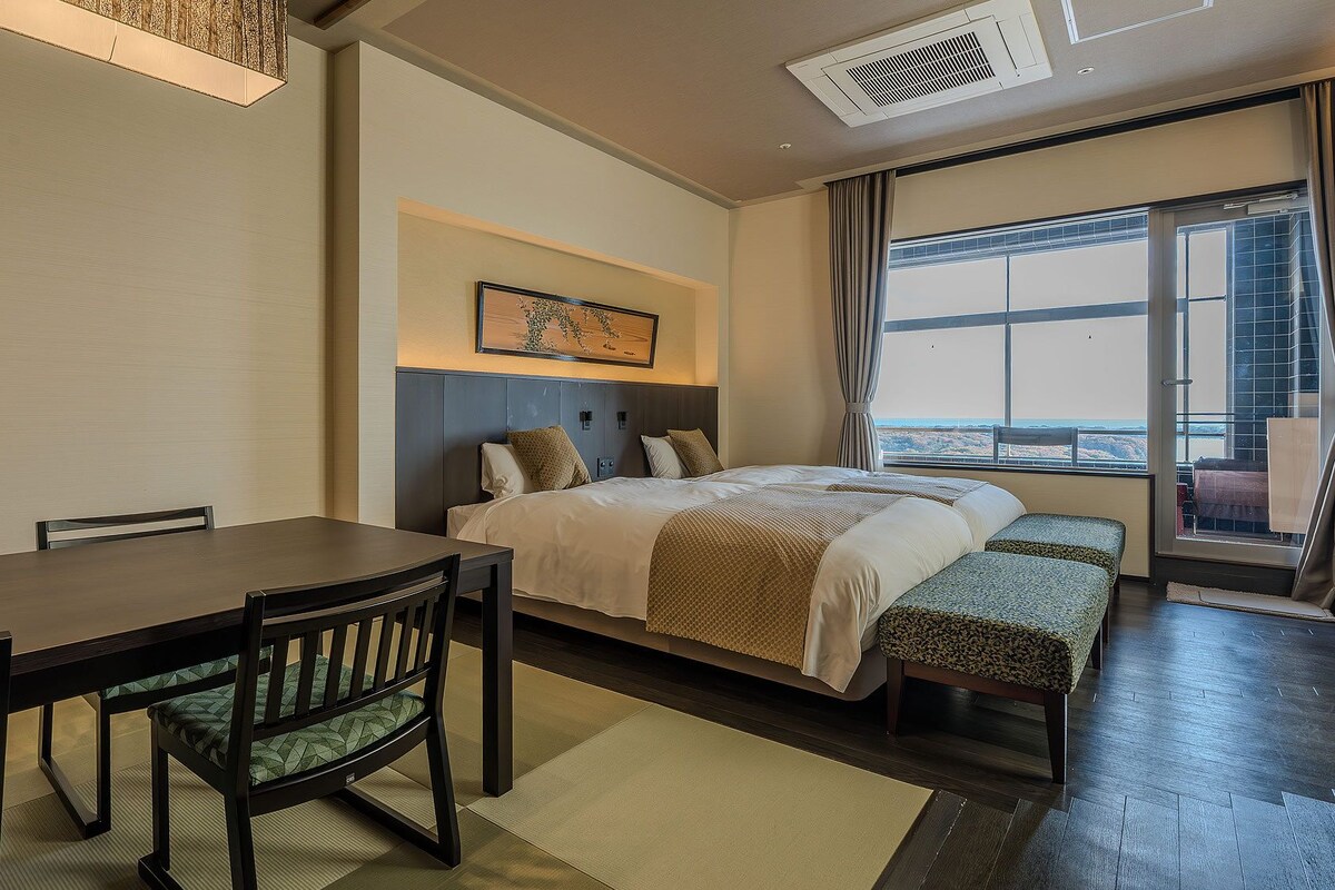Una habitación de hotel con una cama, una mesa de comedor con sillas, un banco, un cuadro en la pared y un ventanal con vistas.