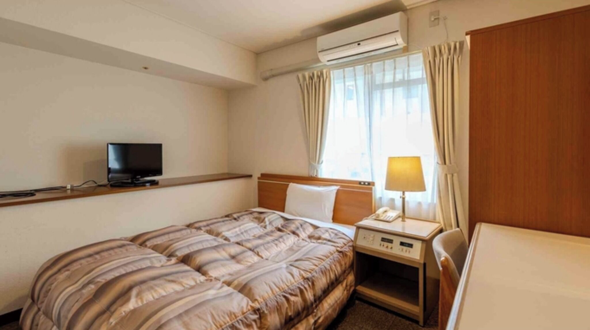 Een hotelkamer met een bed, nachtkastje met lamp, televisie en airconditioning.