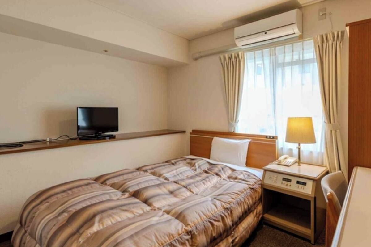 A hotel room with a bed, a bedside table with a lamp, a TV, and an air conditioning unit.
