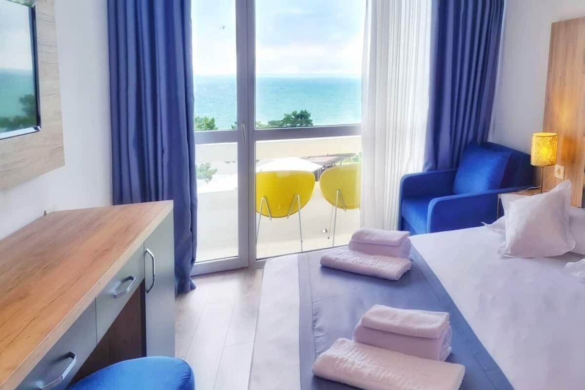 Habitación de hotel con cama, escritorio, silla y vista al mar, además de un balcón con sillas amarillas.