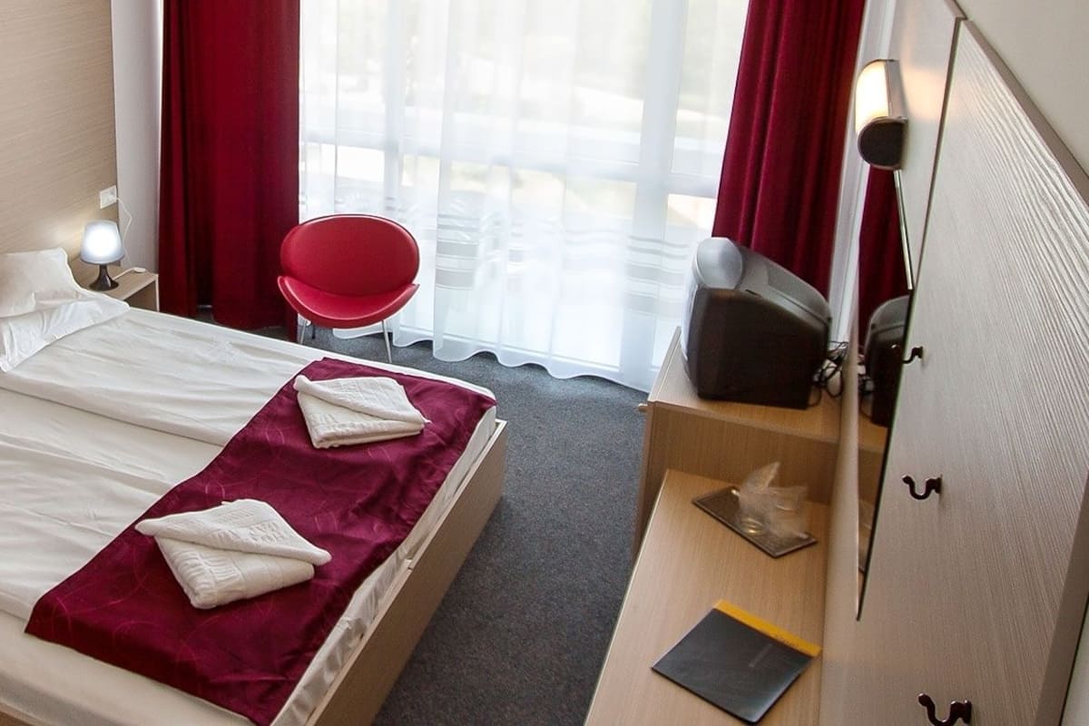 Una habitación de hotel con una cama, dos mesitas de noche, una silla roja, un escritorio y un espejo.