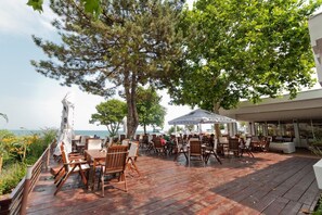 An outdoor patio with wooden tables and chairs, a white umbrella, and a view of the sea. - HOTEL OPAL Cap Aurora (Jupiter)