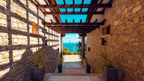 Un pasillo de piedra con una pérgola de madera, plantas en macetas y vistas al mar.