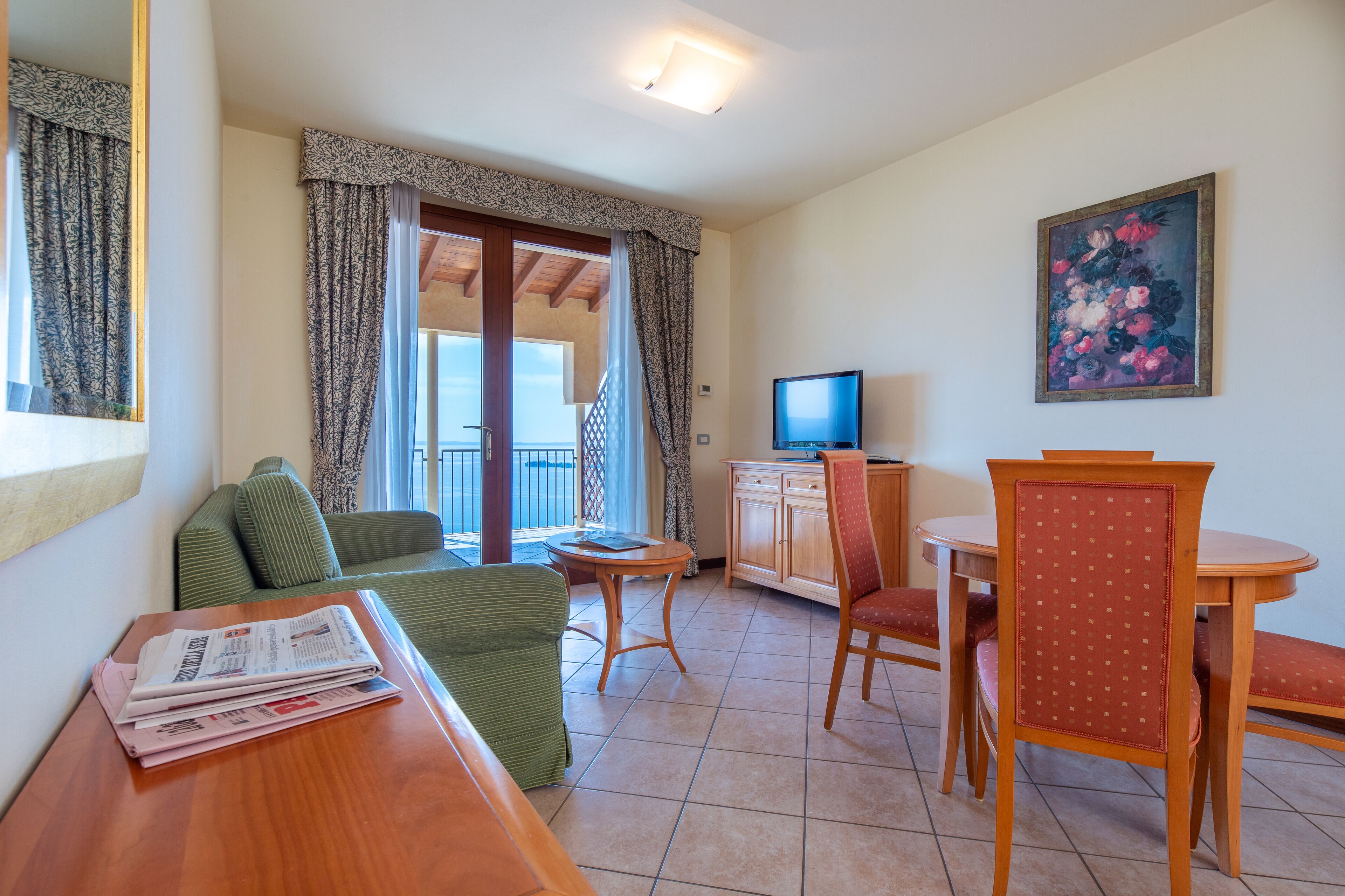 A hotel room with a balcony, a dining area, a TV, and a painting on the wall.