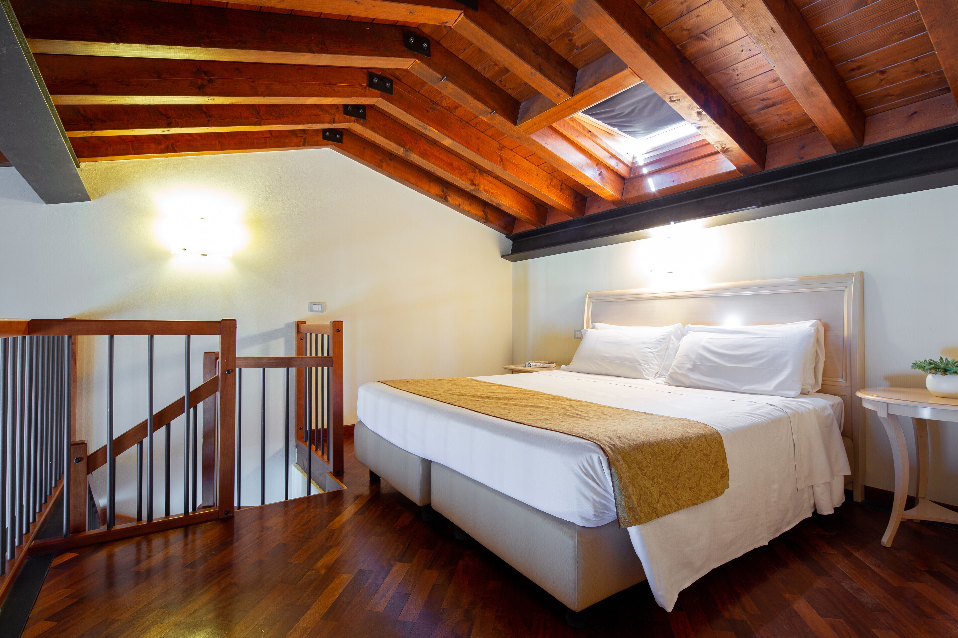 A bedroom with a wooden ceiling, a bed with white bedding, a small bedside table, and a staircase with a wooden railing.