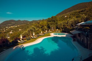 A resort pool with lounge chairs and umbrellas, surrounded by lush greenery and mountains. - Domina Borgo degli Ulivi - Lake Garda (Gardone Riviera)